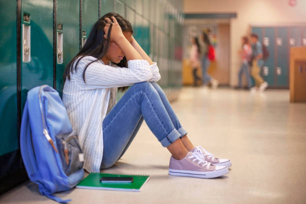 Frustrated teenage female student sitting with head in hands. Side view of high school girl in illuminated corridor. She is against metallic lockers.